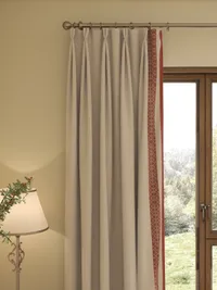 Beige curtain with orange pattern border hanging next to a wooden window and a lamp